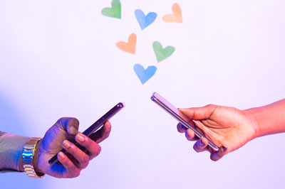 two hands, each holding a cell phone, with artistic hearts floating up between them
